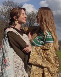 two women standing next to each other in front of a field with trees and clouds