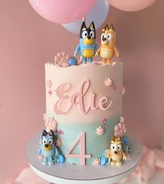 a pink and blue cake with cartoon characters on it's top, surrounded by balloons