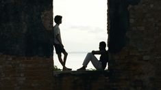 two people sitting on the edge of an old brick building looking out at water and sky