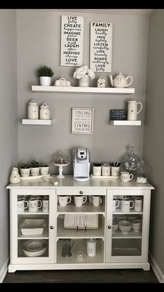 there is a white buffet with dishes and cups on the top shelf, along with two framed pictures above it