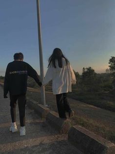 two people holding hands while walking down the street at sunset or dawn with trees in the background