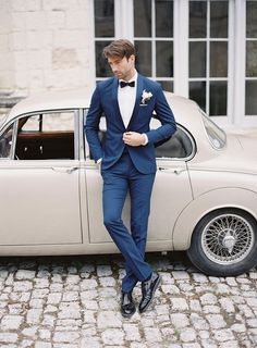 a man in a tuxedo leaning against a car on a cobblestone street