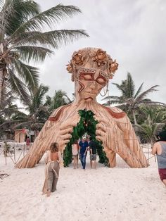 people standing in front of a large wooden sculpture on the beach with palm trees behind them