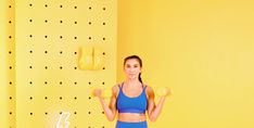 a woman standing in front of a yellow wall with her hands up and holding two dumbs