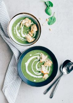 two bowls of green soup with croutons and spinach leaves on the side