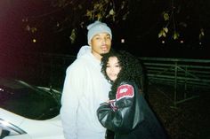 a man and woman standing next to each other in front of a car at night