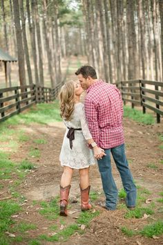 a man and woman kissing in the middle of a path surrounded by pine trees on a farm