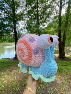 a hand holding up a stuffed animal in front of some trees and water, with the image of a snail on it's back