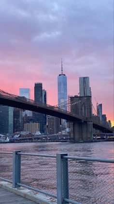 a bridge that is over some water with buildings in the back ground and a pink sky