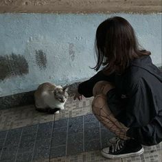 a woman kneeling down next to a white and gray cat on a tiled floor in front of a blue wall