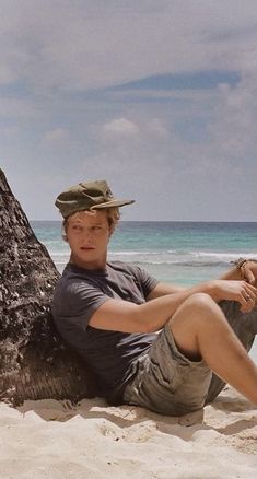 a man sitting on top of a sandy beach next to the ocean with a tree trunk in front of him