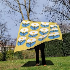 a person holding up a yellow and blue quilt with flowers on it in the grass