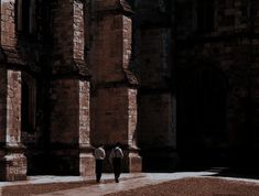 two people are walking in front of an old building with stone pillars and arches on either side