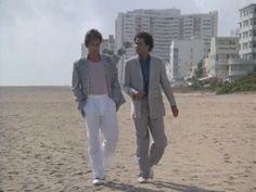 two men walking on the beach with buildings in the background