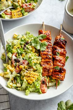 two plates filled with chicken, salad and corn on the cob skewers