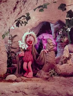 the muppet is holding flowers in his hand and standing next to a stone cave