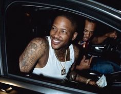 two men are sitting in the back seat of a car and one is wearing a white tank top