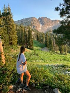 a woman standing on top of a lush green hillside next to a forest filled with lots of trees