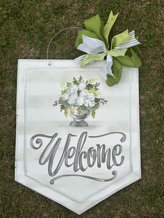 a welcome sign on the grass with flowers in a vase hanging from it's side