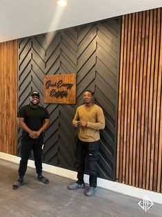 two men standing next to each other in front of a wall with wood paneling