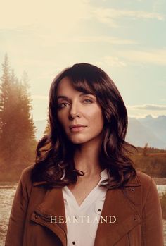 a woman standing in front of a lake wearing a brown jacket and white shirt with the words heartland on it