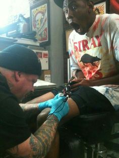 a man getting his arm tattoo done by another man in a barbershop at the same time