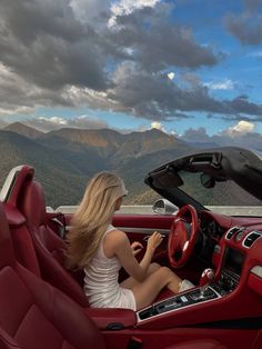 a woman sitting in the driver's seat of a red sports car with mountains in the background