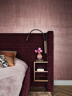 a bed with a purple headboard next to a pink flower on a shelf and a lamp