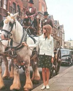 a woman standing next to a horse drawn carriage on a city street with two men riding in the back