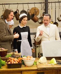 three people standing in front of a table full of vegetables and fruit, with one person wearing a white apron