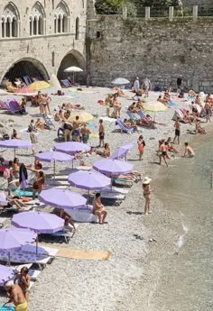 many people are on the beach with purple umbrellas and blue chairs in front of an old stone building