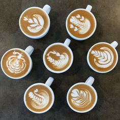 six cups of latte art coffee with designs on the top and bottom, sitting on a table