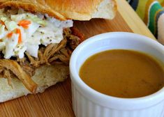 a pulled pork sandwich with coleslaw and a cup of gravy on a cutting board