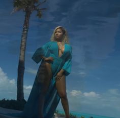 a woman in a blue dress standing next to a palm tree on the ocean shore