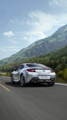 the rear end of a silver sports car driving down a road with mountains in the background