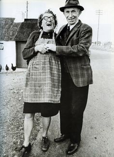 an old man and woman standing next to each other in front of a farm house