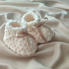 a pair of white crocheted baby booties with bows on the side laying on a blanket