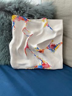 a white purse with colorful paint splattered on it sitting on a blue bed