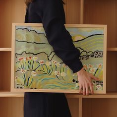 a woman holding up a painting in front of a book shelf filled with wooden shelves