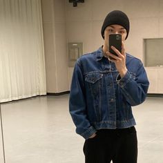 a man in a blue jean jacket taking a selfie with his cell phone while wearing black pants and a beanie