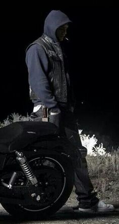 a man standing next to a motorcycle in the dark with his hood up and boots on