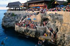 many people are standing on the edge of a cliff by the ocean while others watch