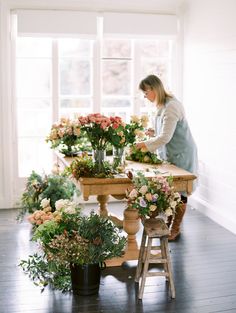 a woman arranging flowers on a table in a room with wooden floors and white walls