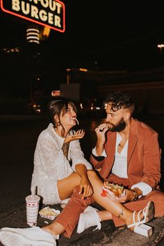 a man and woman sitting on the ground eating food at a fast food restaurant with a neon sign in the background