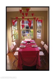 the table is set for valentine's day with paper hearts hanging from the ceiling