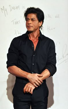 a man in black shirt and pants standing next to a white wall with writing on it