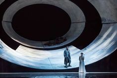 two people standing in front of a large circular object on a stage with water surrounding them