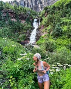 a woman standing in front of a waterfall with trees and bushes around her, looking up at the falls