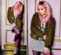 a woman with pink hair wearing plaid pants and a furry jacket is posing in front of a wall