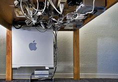 an apple computer under a desk with many wires and plugs attached to the back